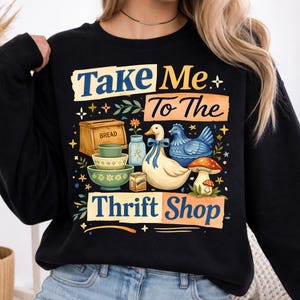 May include: Black sweatshirt with the text "Take Me To The Thrift Shop" and illustrations of a duck, chicken, mushroom, and bowls. The design includes a bread box and a jar. The sweatshirt is a cozy, casual style.