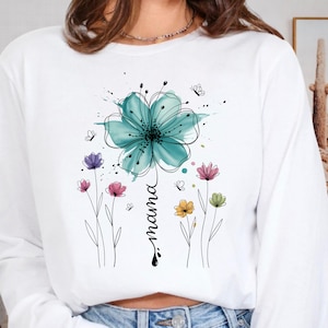 May include: White long-sleeve sweatshirt featuring a watercolor floral design. A large teal flower is at the top, with the word "mama" written vertically below it. Smaller colorful flowers and butterflies are also present.