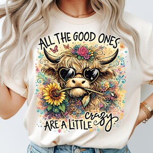 May include: A cream-colored t-shirt featuring a graphic of a Highland cow wearing heart-shaped sunglasses and holding a sunflower in its mouth. The design includes colorful floral accents, butterflies, and the text ALL THE GOOD ONES ARE A LITTLE crazy.
