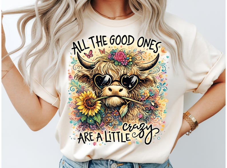 May include: A cream-colored t-shirt featuring a graphic of a Highland cow wearing heart-shaped sunglasses and holding a sunflower in its mouth. The design includes colorful floral accents, butterflies, and the text ALL THE GOOD ONES ARE A LITTLE crazy.