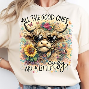 May include: A cream-colored t-shirt featuring a graphic of a shaggy highland cow wearing heart-shaped sunglasses and holding a sunflower in its mouth. The design includes colorful floral accents, butterflies, and the text ALL THE GOOD ONES ARE A LITTLE CRAZY.
