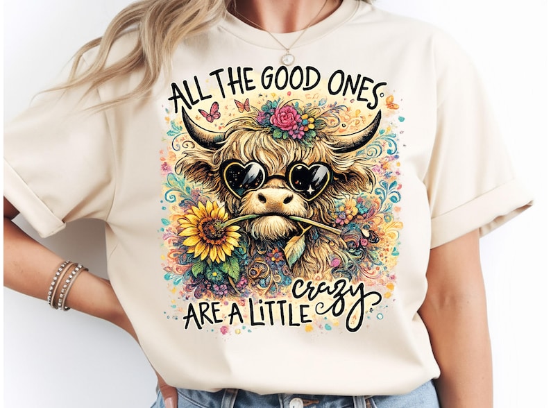 May include: A cream-colored t-shirt featuring a graphic of a shaggy highland cow wearing heart-shaped sunglasses and holding a sunflower in its mouth. The design includes colorful floral accents, butterflies, and the text ALL THE GOOD ONES ARE A LITTLE CRAZY.