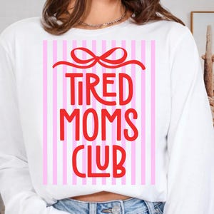 May include: White long-sleeve sweatshirt with a red bow and the words "TIRED MOMS CLUB" in red block letters. The background features pink and white vertical stripes. A casual and comfortable top.