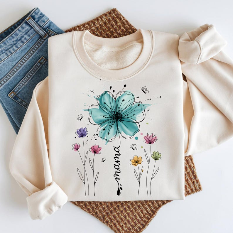 May include: Cream-colored sweatshirt featuring a large teal watercolor flower with black outlines and the word "mama" in a vertical script. Additional colorful flowers and butterflies are scattered around the design. The sweatshirt is folded with a pair of blue jeans.