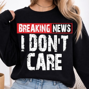 Puede incluir: Una sudadera negra de manga larga con un diseño gráfico que presenta el texto BREAKING NEWS en letras blancas sobre un fondo rectangular rojo, seguido del texto I DON'T CARE en letras mayúsculas blancas grandes y desgastadas.