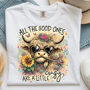 May include: A white t-shirt featuring a colorful graphic of a Highland cow wearing heart-shaped sunglasses and holding a sunflower in its mouth. The text reads ALL THE GOOD ONES ARE A LITTLE crazy, surrounded by floral and butterfly illustrations.