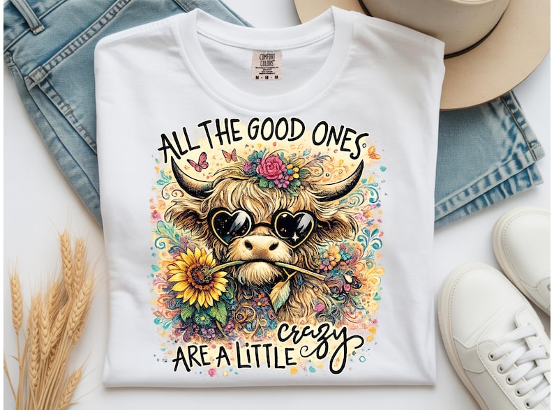 May include: A white t-shirt featuring a colorful graphic of a Highland cow wearing heart-shaped sunglasses and holding a sunflower in its mouth. The text reads ALL THE GOOD ONES ARE A LITTLE crazy, surrounded by floral and butterfly illustrations.