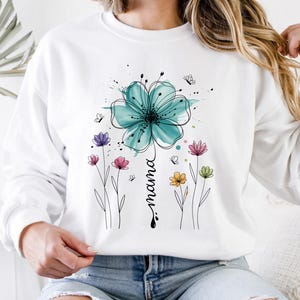 May include: White sweatshirt featuring a large teal watercolor flower with black outlines and the word "mama" in a script font. Additional colorful flowers and butterflies are also depicted, creating a floral design.