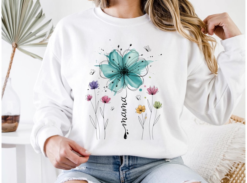 May include: White sweatshirt featuring a large teal watercolor flower with black outlines and the word "mama" in a script font. Additional colorful flowers and butterflies are also depicted, creating a floral design.