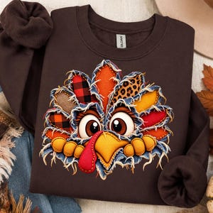 May include: A dark brown crewneck sweatshirt featuring a central graphic of a turkey face. The turkey is composed of various fabric-like patches including red and black buffalo plaid, leopard print, and denim textures with frayed edges.