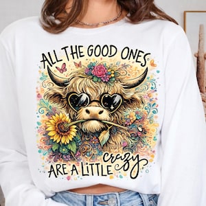 May include: A white long-sleeve sweatshirt featuring a graphic of a Highland cow wearing heart-shaped sunglasses and holding a sunflower in its mouth. The design includes colorful floral patterns, butterflies, and the text ALL THE GOOD ONES ARE A LITTLE crazy.