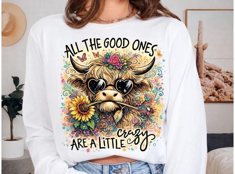 May include: A white long-sleeve sweatshirt featuring a graphic of a Highland cow wearing heart-shaped sunglasses and holding a sunflower in its mouth. The design includes colorful floral patterns, butterflies, and the text ALL THE GOOD ONES ARE A LITTLE crazy.