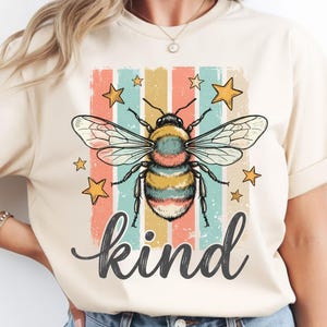 May include: Cream-colored t-shirt featuring a colorful bee illustration with the word "kind" in a cursive font. The bee is surrounded by stars and set against a retro striped background.