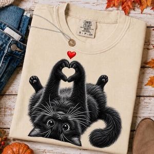 Może przedstawiać: Beżowy t-shirt z krótkim rękawem z grafiką czarnego kota leżącego na plecach. Kot ma duże oczy i trzyma przednie łapy razem, tworząc kształt serca, z małą czerwoną ikoną serca umieszczoną nad łapami.