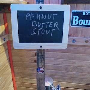 May include: A wooden sign with the words "PEANUT BUTTER STOUT" in white chalk. The sign is mounted on a wooden post with an American flag sticker. The background is a wooden wall.