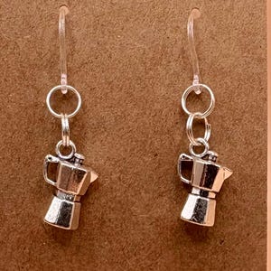 Może przedstawiać: Srebrne kolczyki w kształcie ekspresu do kawy wiszące na przezroczystych plastikowych haczykach. Kolczyki są prezentowane na brązowej karcie z otworem u góry. Tło jest w jednolitym kolorze brzoskwiniowym.