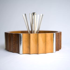 Puede incluir: Un portautensilios de madera con un diseño geométrico. El soporte está hecho de madera clara y oscura con detalles metálicos plateados. Los utensilios plateados están dispuestos en el centro.