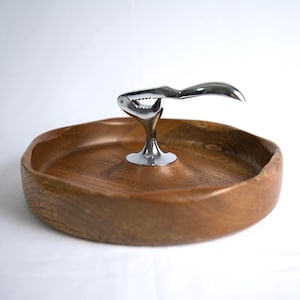 Puede incluir: Una bandeja de madera para servir con un cascanueces cromado incorporado. La bandeja es redonda con un acabado de veta de madera natural. El cascanueces está montado en el centro y tiene un diseño de palanca.