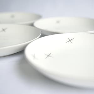 May include: Four white ceramic plates with a minimalist design. Each plate features a simple gray cross-shaped accent. The plates are arranged on a white surface, with a shallow depth of field.