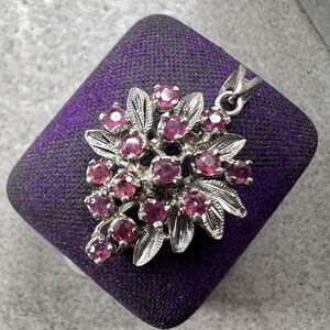 May include: A silver pendant featuring a cluster of small, round, deep pink gemstones arranged among textured silver leaves. The pendant includes a metal bail at the top for attachment to a chain, showcasing a detailed floral-inspired design.