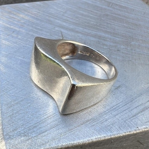 Könnte beinhalten: Ein silberfarbener Ring mit einer breiten, glatten und geschwungenen Ringschiene, die einen markanten, erhöhten und kantigen oberen Bereich aufweist. Die Metalloberfläche ist poliert mit leichten Gebrauchsspuren und sichtbaren Punzierungen.