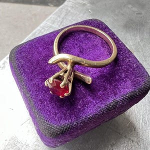 May include: A gold ring featuring a single round-cut red gemstone set in a four-prong mounting. The band has a smooth, polished finish and is positioned on a square, deep purple velvet cushion with dark trim.