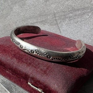 May include: A silver cuff bracelet featuring a repeating stamped pattern of sunbursts and small circular motifs along the outer surface. The metal has a polished finish with darkened recessed areas that highlight the intricate stamped designs.