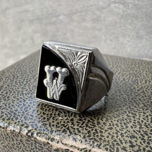 May include: A silver-toned ring featuring a trapezoidal face with a black inlay. The center of the black inlay displays a raised, metallic letter W. The metal surrounding the inlay is decorated with engraved floral and geometric patterns.