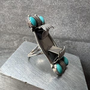 Pode incluir: Um anel longo de prata com dois conjuntos de cabochões de turquesa ovais na parte superior e inferior. A banda de prata tem um design curvo e alongado com detalhes metálicos decorativos em forma de folha que rodeiam as pedras azul-brilhantes.