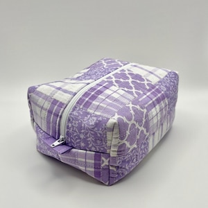 May include: A quilted, rectangular cosmetic bag with a white zipper. The bag features a patchwork design in shades of lavender and white, with floral, plaid, and geometric patterns. The bag is designed to hold makeup and other beauty products.