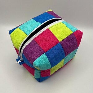 May include: A rectangular fabric pouch featuring a patchwork design of square panels in bright shades of lime green, turquoise, royal blue, magenta, and purple. The pouch includes a white zipper closure along the top edge with a white pull tab.