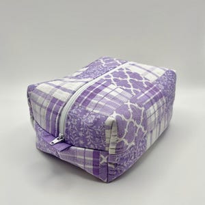 May include: A rectangular quilted pouch featuring a patchwork design in shades of lavender, purple, and white. The fabric displays various patterns including floral motifs, plaid, and geometric lattice shapes. A white zipper runs along the top.