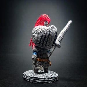 Puede incluir: Una figura de caballero tejida a ganchillo sobre una base circular. El caballero lleva una armadura gris con un penacho rojo en el casco, detalles azules en los brazos y un cinturón naranja. La figura sostiene una lanza gris larga y puntiaguda.