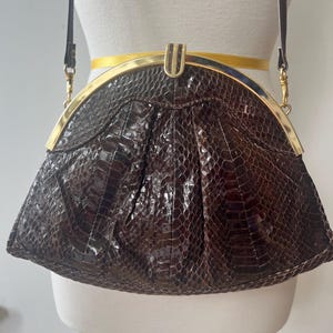 May include: A brown, textured handbag with a gold-colored clasp and trim. The bag has a shoulder strap and a curved shape. The bag is made of a material that resembles snakeskin.