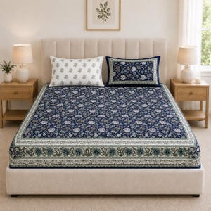 Könnte beinhalten: Ein Bett mit einer Matratze, die mit einem dunkelblauen Laken mit weißem und grünem Blumenmuster bezogen ist, umrandet von einem breiten Band mit komplexen botanischen Motiven. Zwei rechteckige Kissen liegen am Kopfende, eines weiß mit grauen Drucken.