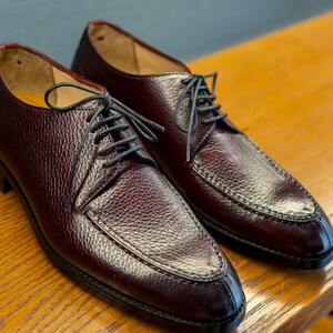 May include: A pair of burgundy leather dress shoes with black laces. The shoes have a textured surface and a classic design, suitable for formal occasions. The shoes are displayed on a wooden surface.