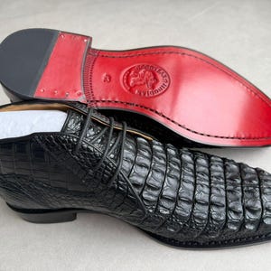 May include: Black leather ankle boots with a crocodile texture. The sole is red with a black heel. The boots are laced up and appear to be new.