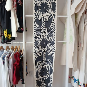 Peut inclure: Une longue écharpe textile crème avec un motif noir répété d'oiseaux, de fleurs et de cerfs. La nappe est drapée sur une étagère blanche en forme d'échelle, avec des vêtements suspendus de chaque côté.