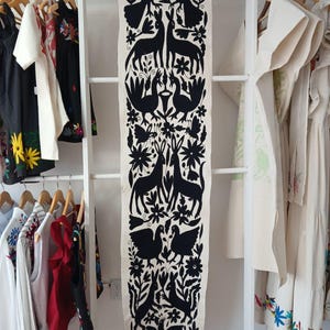 Puede incluir: Un camino de mesa textil color crema con un diseño floral y animal negro. El camino de mesa presenta ciervos, pájaros y flores estilizados. El camino de mesa está colgado en un estante blanco tipo escalera, con ropa visible a ambos lados.