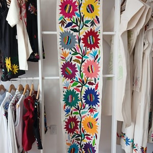 Peut inclure: Une longue nappe rectangulaire en textile blanc cassé avec un motif floral vibrant. Les fleurs brodées sont dans les tons de rose, jaune, bleu et violet, avec des feuilles et des tiges vertes. La nappe est suspendue à un portant blanc, avec des vêtements visibles de chaque côté.