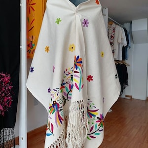 Puede incluir: Poncho color crema con bordados florales y de pájaros coloridos. El poncho presenta un dobladillo con flecos y está adornado con varias flores bordadas en tonos rosa, amarillo, azul y verde. El diseño incluye un motivo de pájaro vibrante.
