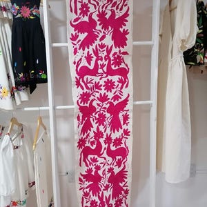 Peut inclure: Une longue pièce de tissu blanche avec un motif répété de cerfs, d'oiseaux et de motifs floraux fuchsia. Le textile est suspendu sur un présentoir blanc en forme d'échelle, mettant en valeur sa broderie complexe et son contraste de couleurs vives.