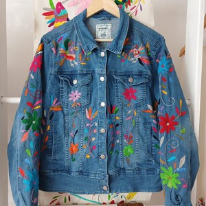 Peut inclure: Veste en jean bleu clair ornée de broderies florales colorées. La veste a une fermeture boutonnée, des poches et des manches longues. La broderie comprend des fleurs et des feuilles rouges, roses, vertes, oranges et bleues. La veste est suspendue à un cintre en bois.