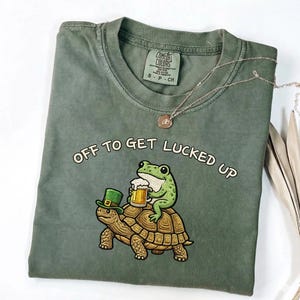 May include: Olive green t-shirt with the phrase "OFF TO GET LUCKED UP" above an illustration of a frog riding a turtle. The frog is holding a beer, and the turtle is wearing a green hat. A gold necklace is draped over the shirt.