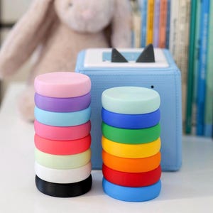 May include: Stacks of colorful silicone rings in various colors, including pink, purple, blue, red, and green. The rings are stacked on a white surface, with a blue box and a stuffed bunny in the background.
