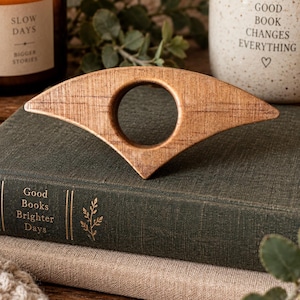 May include: A light brown wooden page holder with a smooth, wing-like shape and a central circular cutout. The object features a visible wood grain texture and a pointed bottom edge, designed to be held between the pages of an open book.