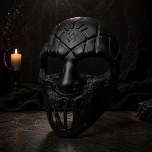 Peut inclure: Un masque noir avec une surface texturée et un design stylisé. Le masque présente un emblème en forme de soleil et un motif géométrique sur le front. La partie inférieure du masque a un design en forme de bouche ouverte et dentelée. Le masque est posé sur un fond sombre.