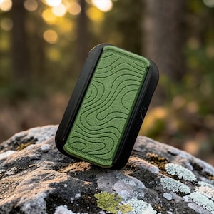 Peut inclure: Un objet rectangulaire noir et vert avec un motif texturé en forme de vagues sur la surface verte. L'objet repose sur une roche moussue, avec un arrière-plan flou de forêt.