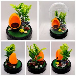 May include: A miniature orange chair with a green plant and a small yellow bird figurine inside a clear glass dome. The base of the dome is black and the scene is set on a green base.