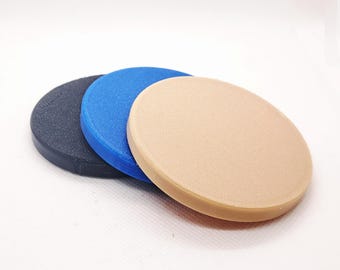 May include: Three round, flat objects in a row. The first is black, the second is blue, and the third is beige. The objects appear to be made of a solid material with a slightly textured surface. The items are arranged on a white background.